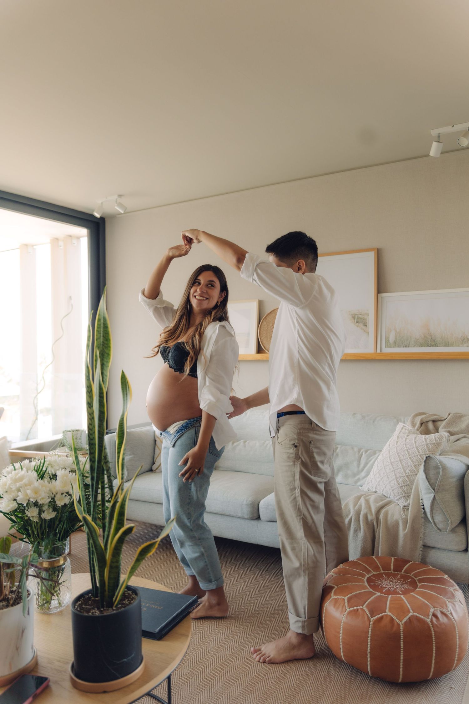 Joyful pregnant couple dancing together in a bright modern living room with plants and neutral decor.