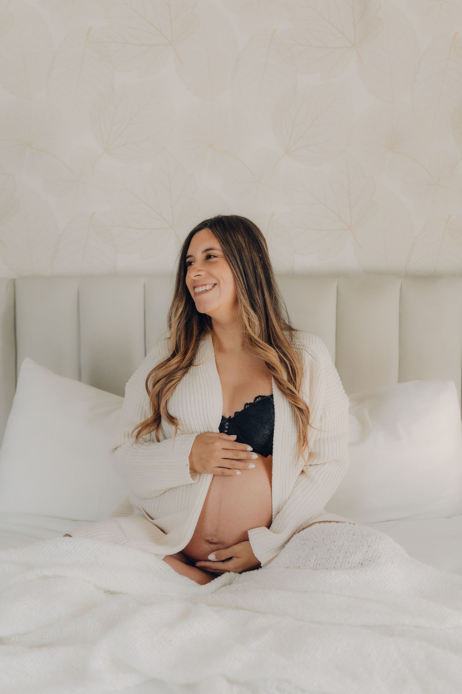 Radiant pregnant woman in cream cardigan and black bra sitting on white bed holding her baby bump.