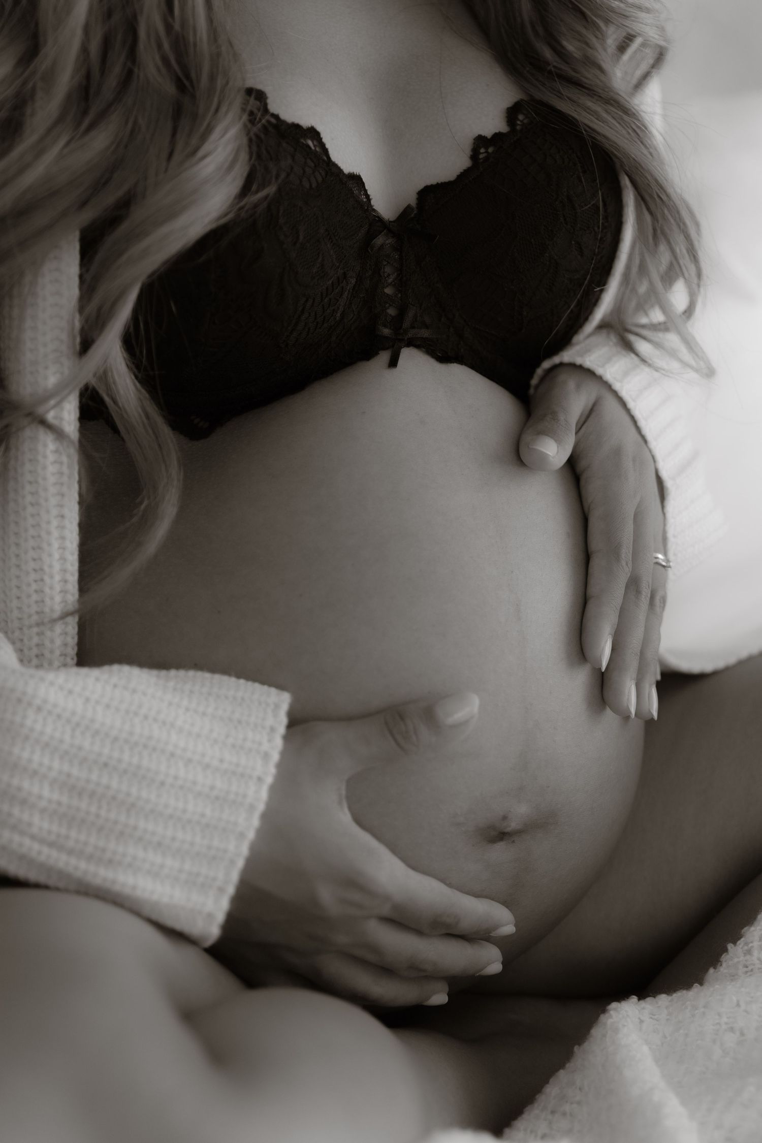 Black and white close-up of pregnant woman in lace bra and knit cardigan cradling her bare bump.