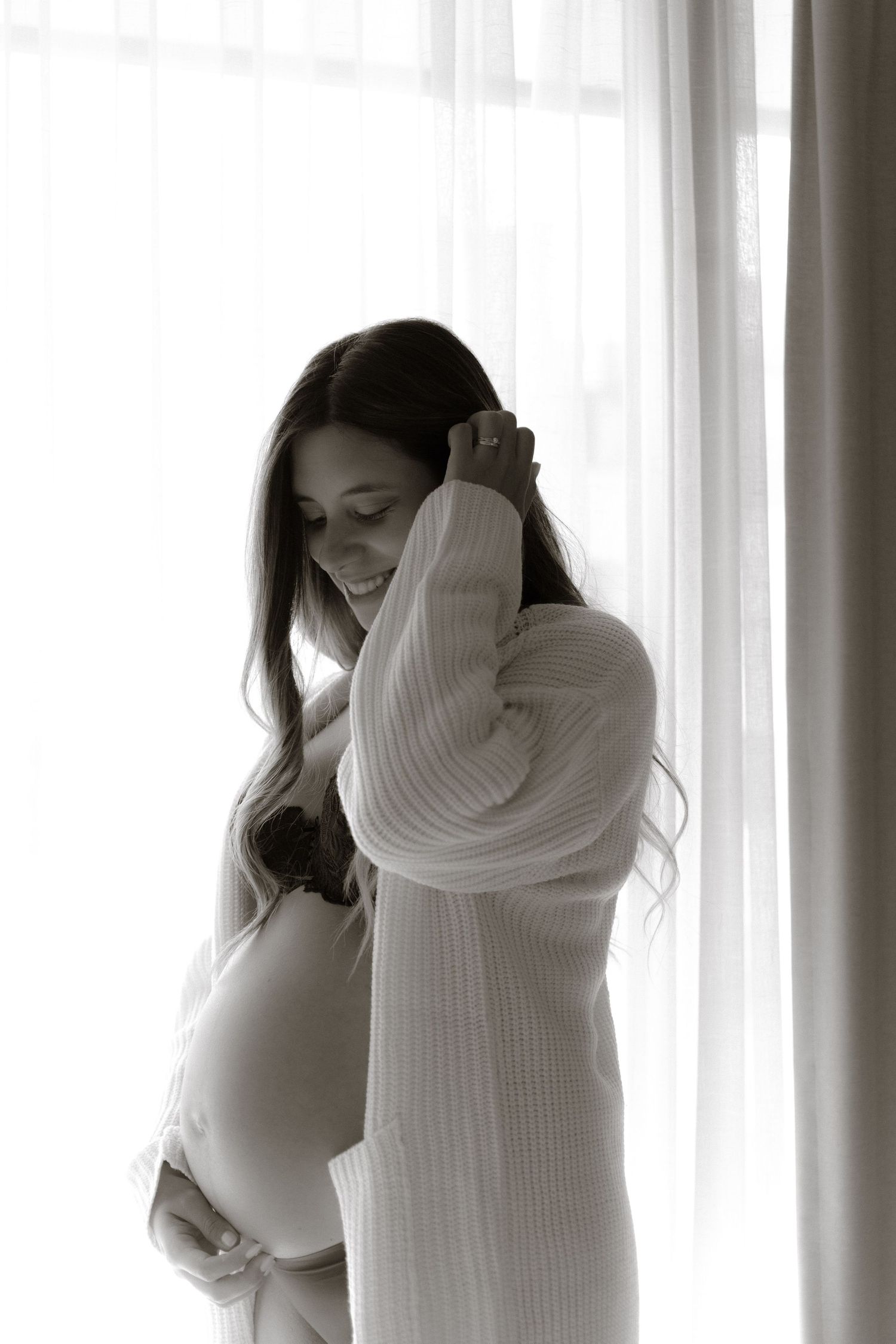 Black and white portrait of pregnant woman in knit cardigan standing by sheer curtained window.