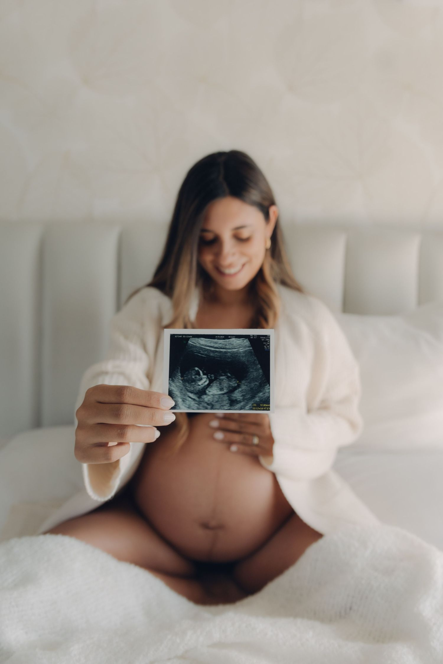 Smiling pregnant woman sitting on bed holding ultrasound photo showing her baby bump in white robe.
