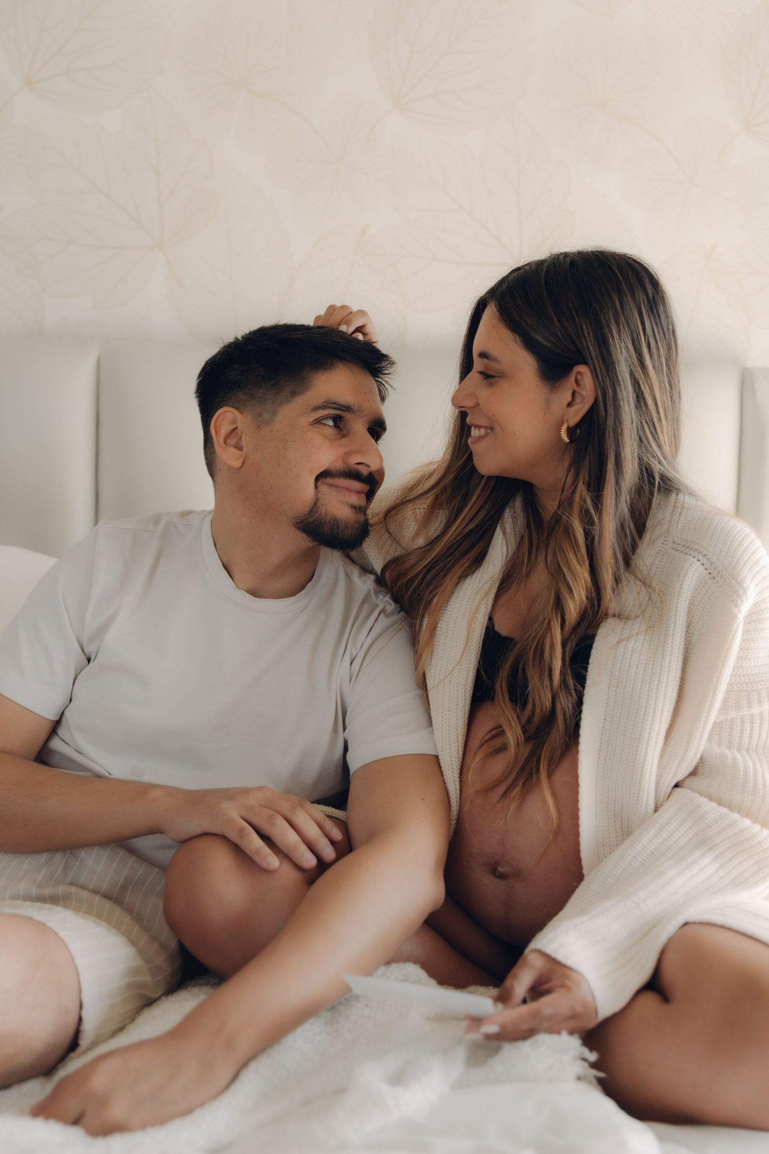 Happy expecting couple sitting on bed smiling at each other, woman in white robe showing baby bump.