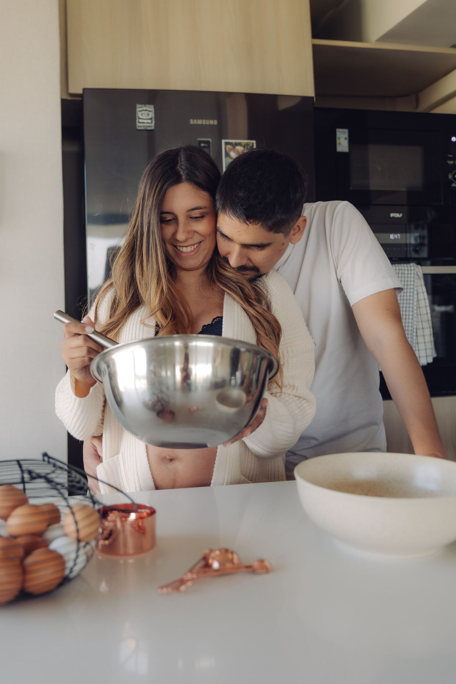 Smiling pregnant couple cooking together in kitchen, woman holding mixing bowl with visible baby bump.
