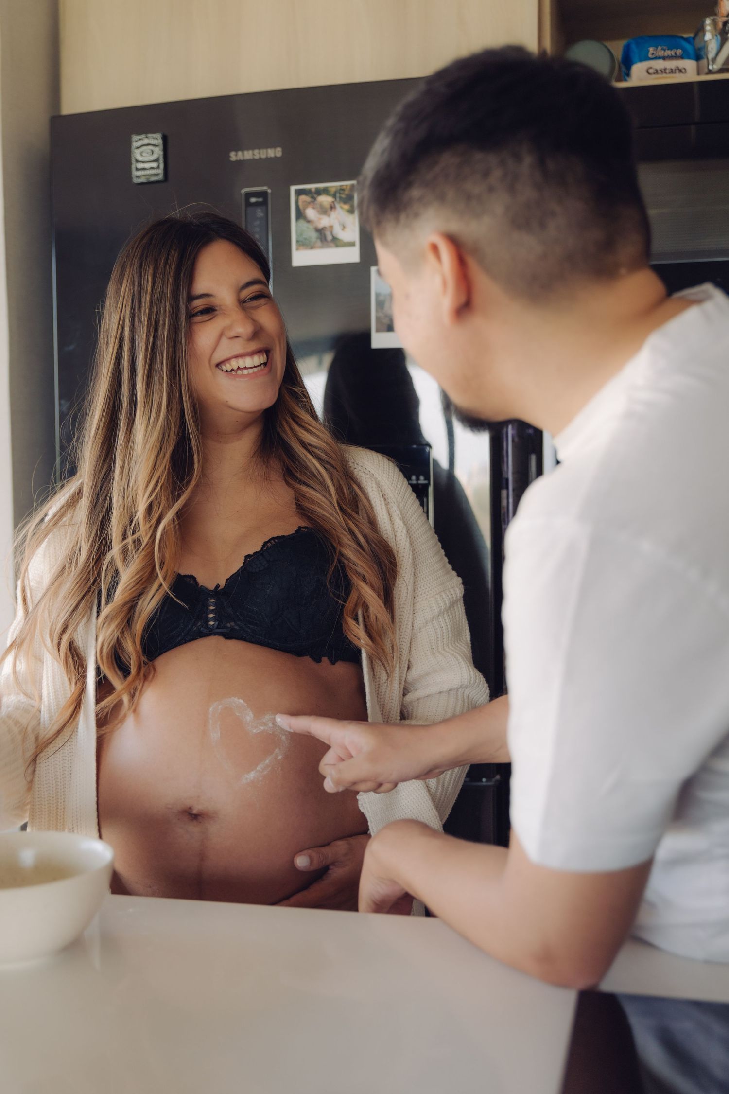 Man playfully drawing heart on laughing pregnant woman's baby bump with cream in kitchen.