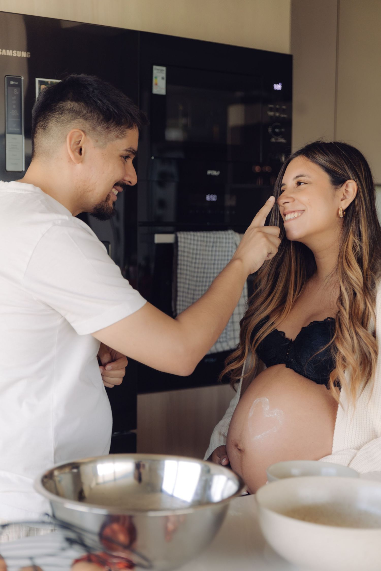 Playful couple in kitchen, man touching pregnant woman's nose with flour while she smiles.