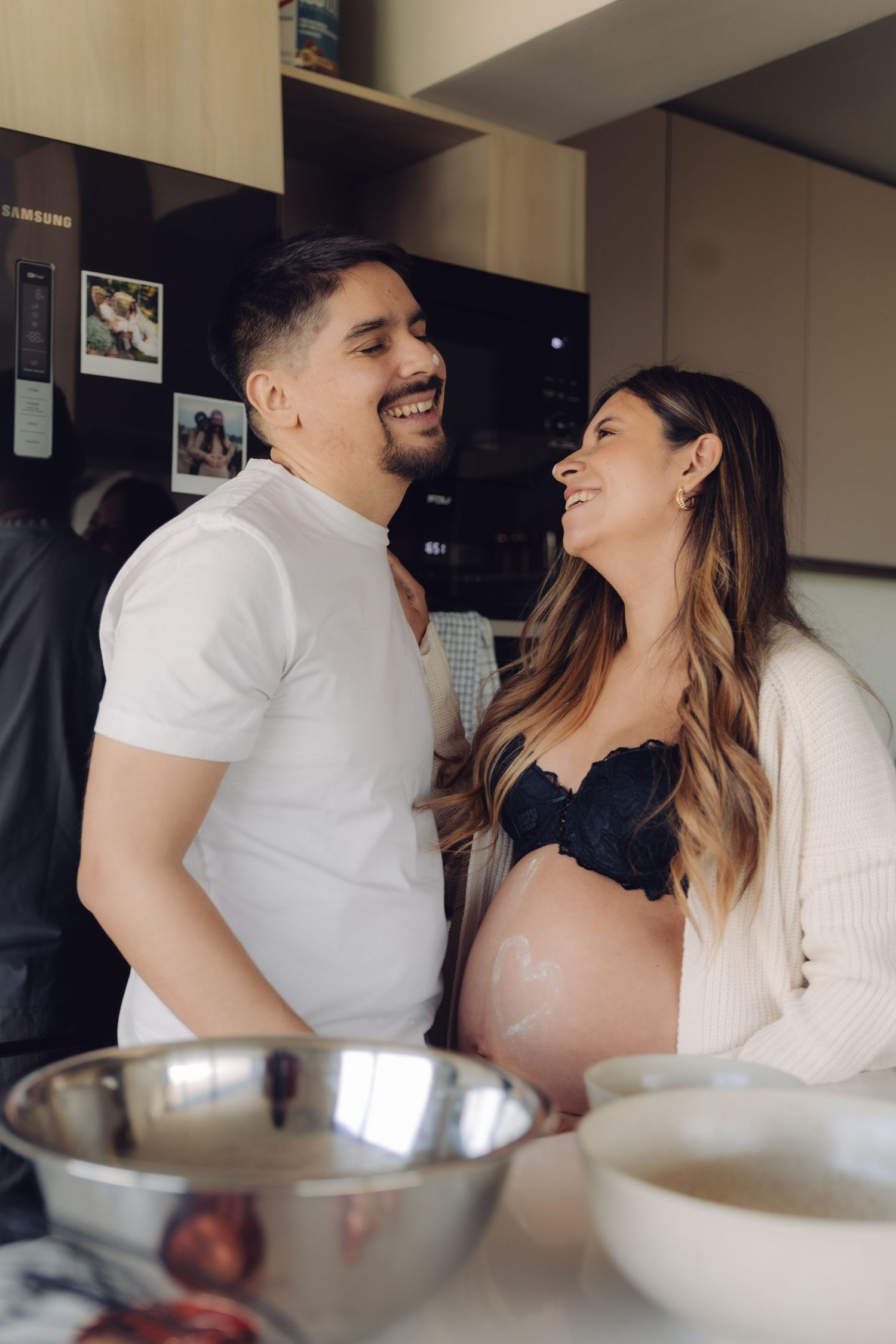 Smiling pregnant couple sharing a joyful moment together in a modern kitchen with mixing bowls on the counter.