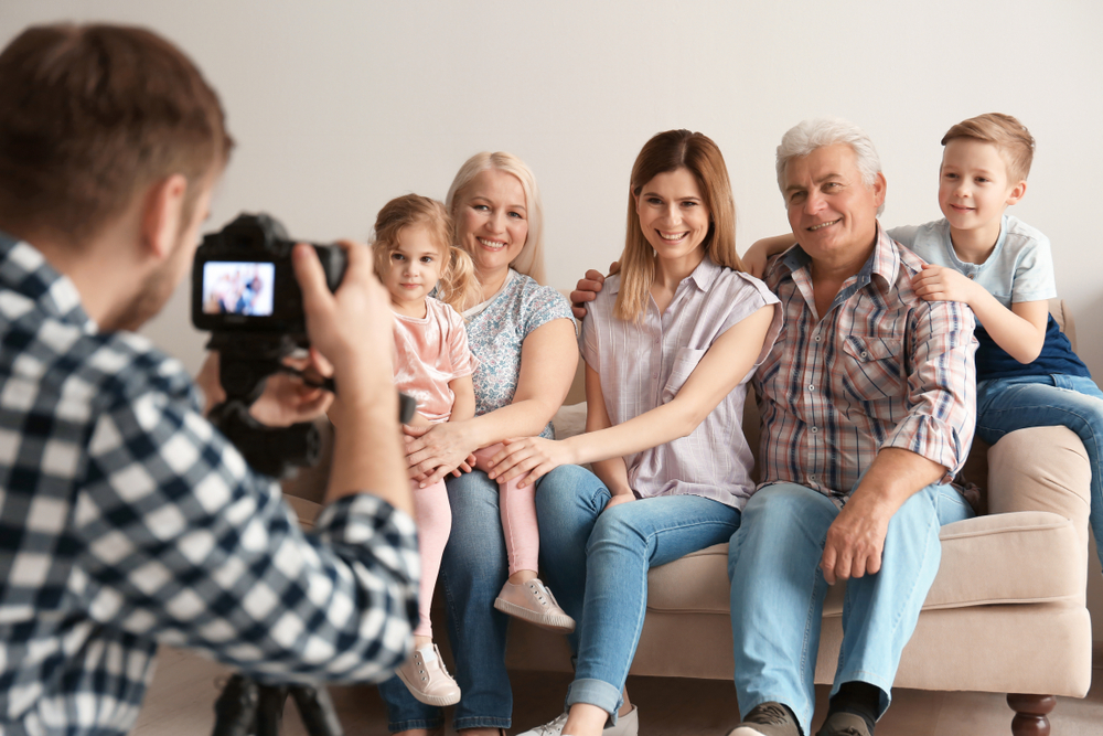 Photographer taking a candid family photo of multi-generational group sitting together on beige couch.