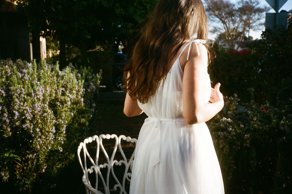 Back view of a person in a flowing white dress standing in a garden near an ornate chair.