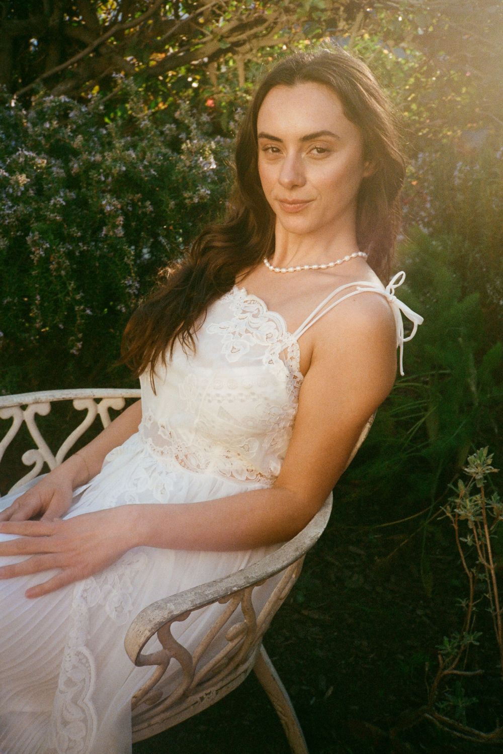 Portrait of a woman in pearl necklace and white lace dress sitting in garden setting.