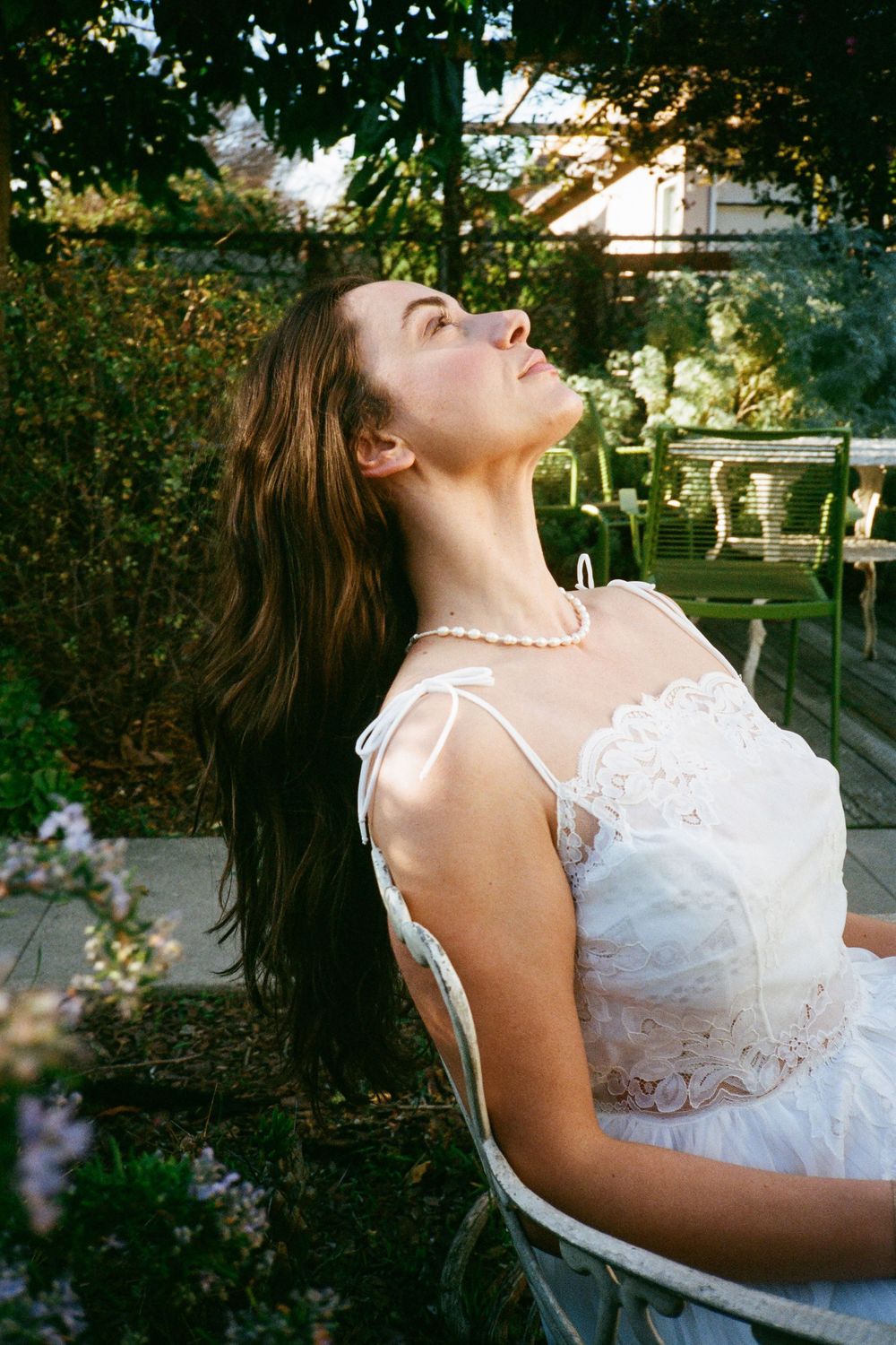 Woman in white lace dress tilts head back while sitting in garden chair during golden hour.