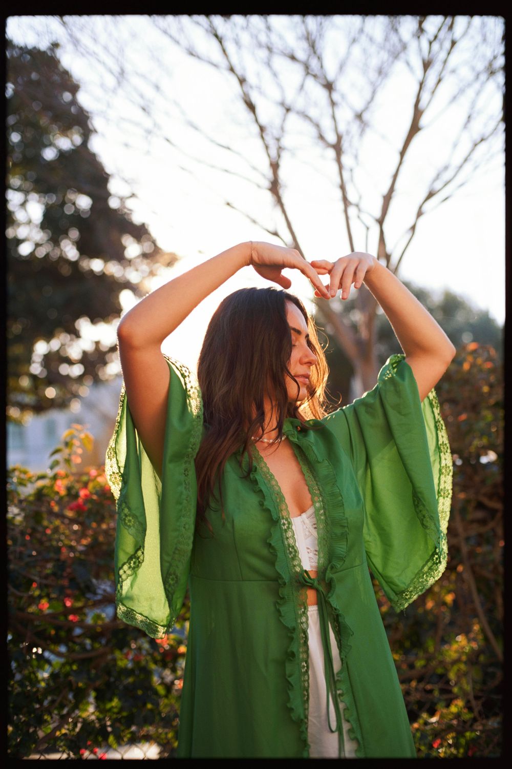 Woman in flowing green dress poses with raised arms in sunlit garden.