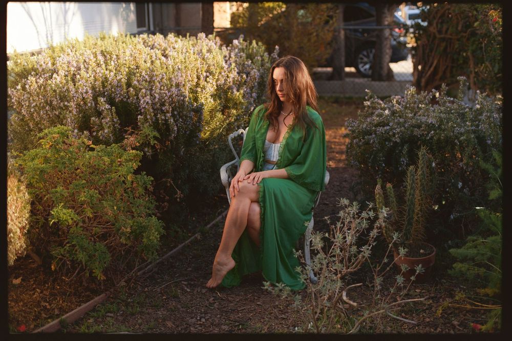 Woman in emerald green dress sitting elegantly in a garden setting at sunset.