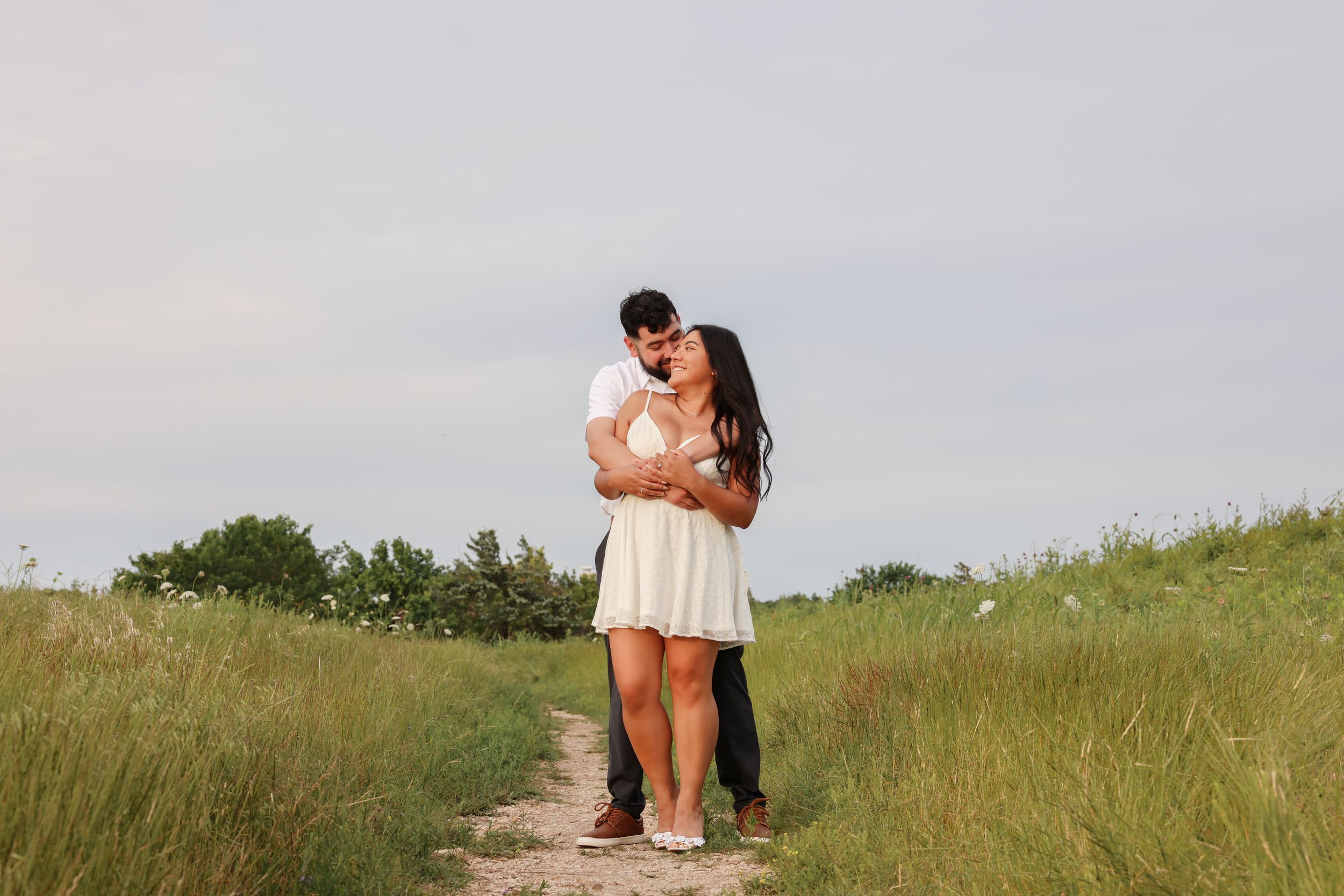 Romantic couple embraces in a golden grassy field during summer sunset creating silhouettes against the sky.