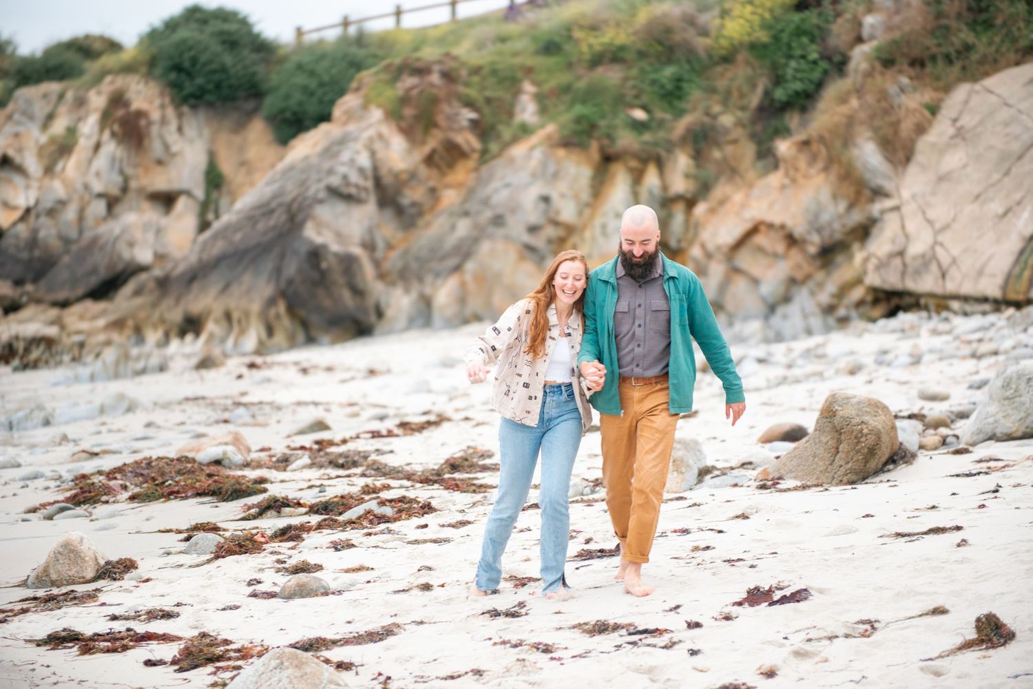 A couple strolls together along a rocky beach with cliffs in the background on an overcast day in monterey.
