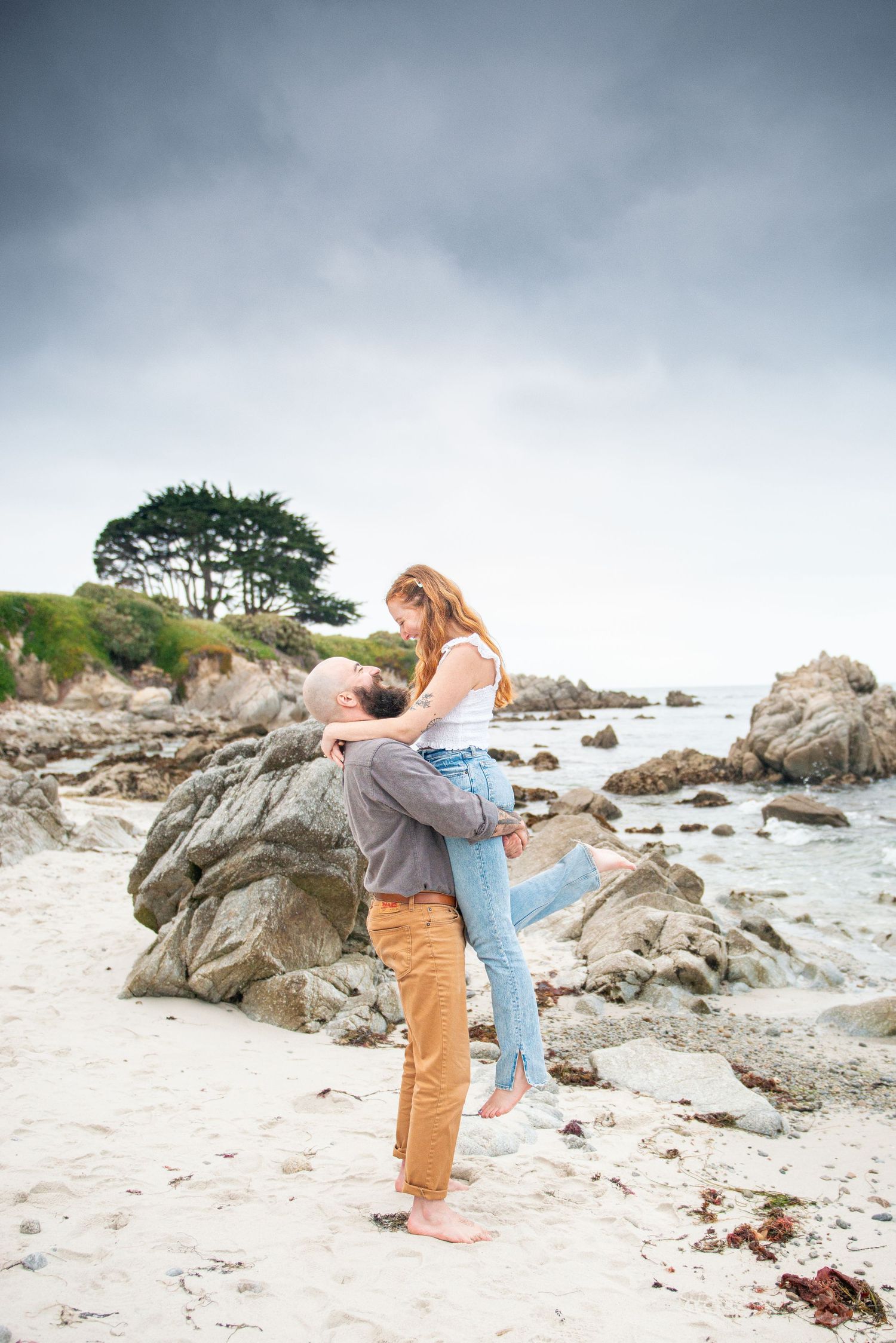 A romantic moment captured on a rocky beach with dramatic stormy skies and crashing waves in the background. in monterey.