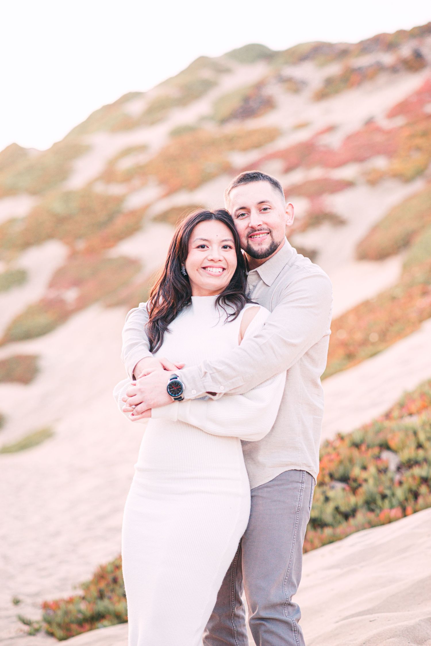 A couple embraces while standing on a sandy hillside with orange coastal vegetation at sunset. in monterey