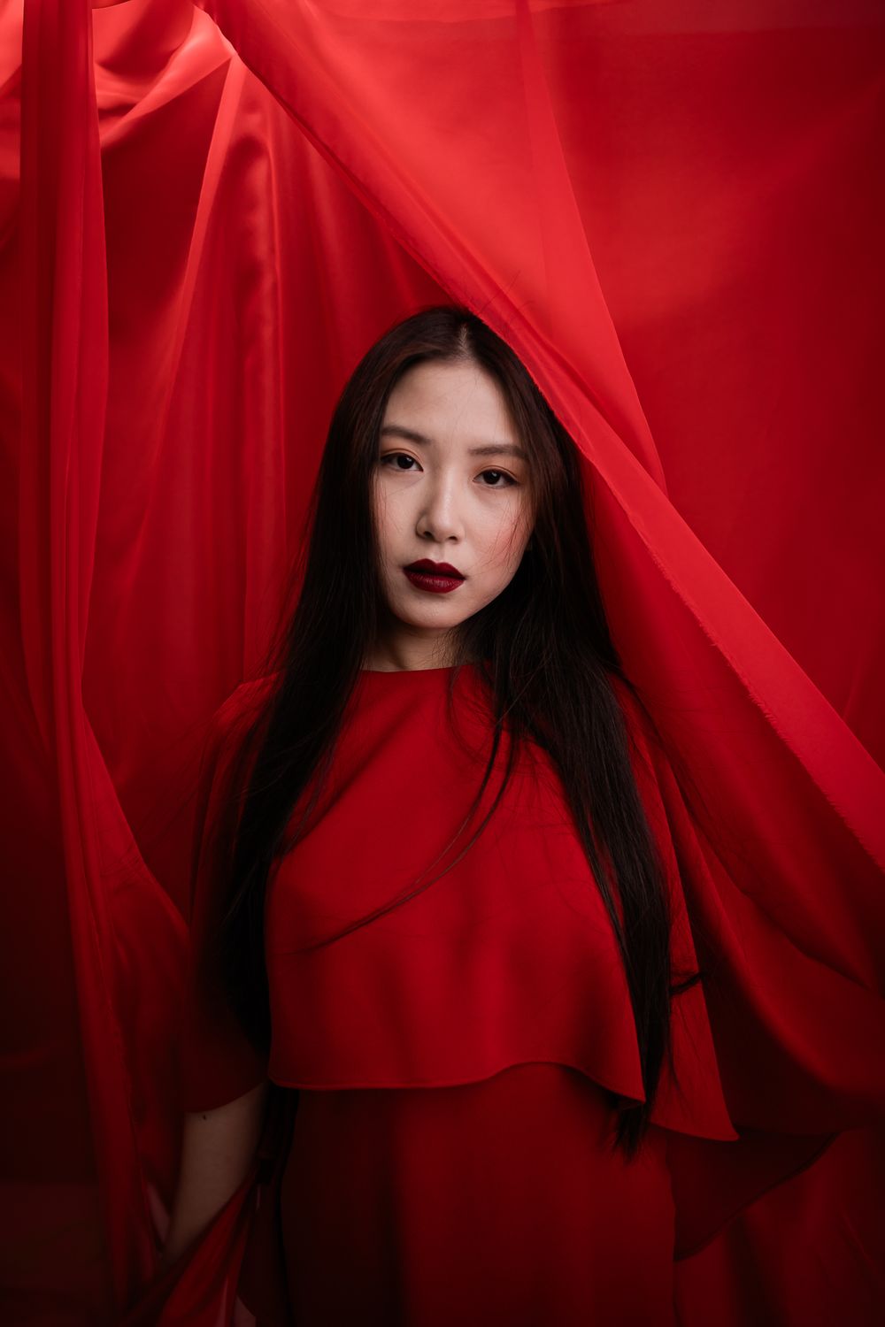 Artistic portrait against flowing red fabric backdrop with dramatic lighting and monochromatic color scheme.