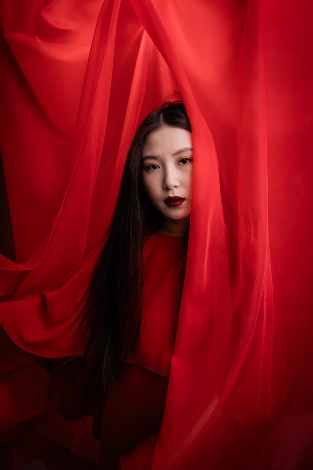 Artistic portrait surrounded by flowing red fabric and drapery against dark background creating dramatic mood.