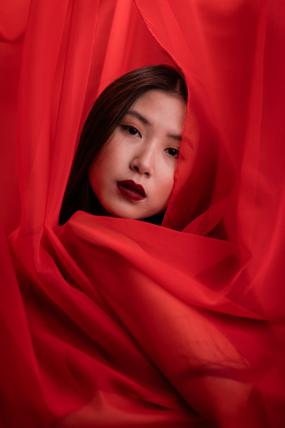 Artistic portrait with vibrant red fabric draping around dark lipstick and moody lighting.