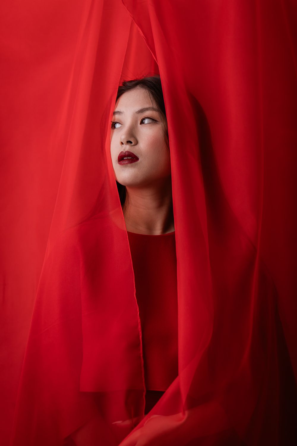 Artistic portrait through red fabric showcasing deep red lipstick and dramatic lighting against vibrant backdrop.
