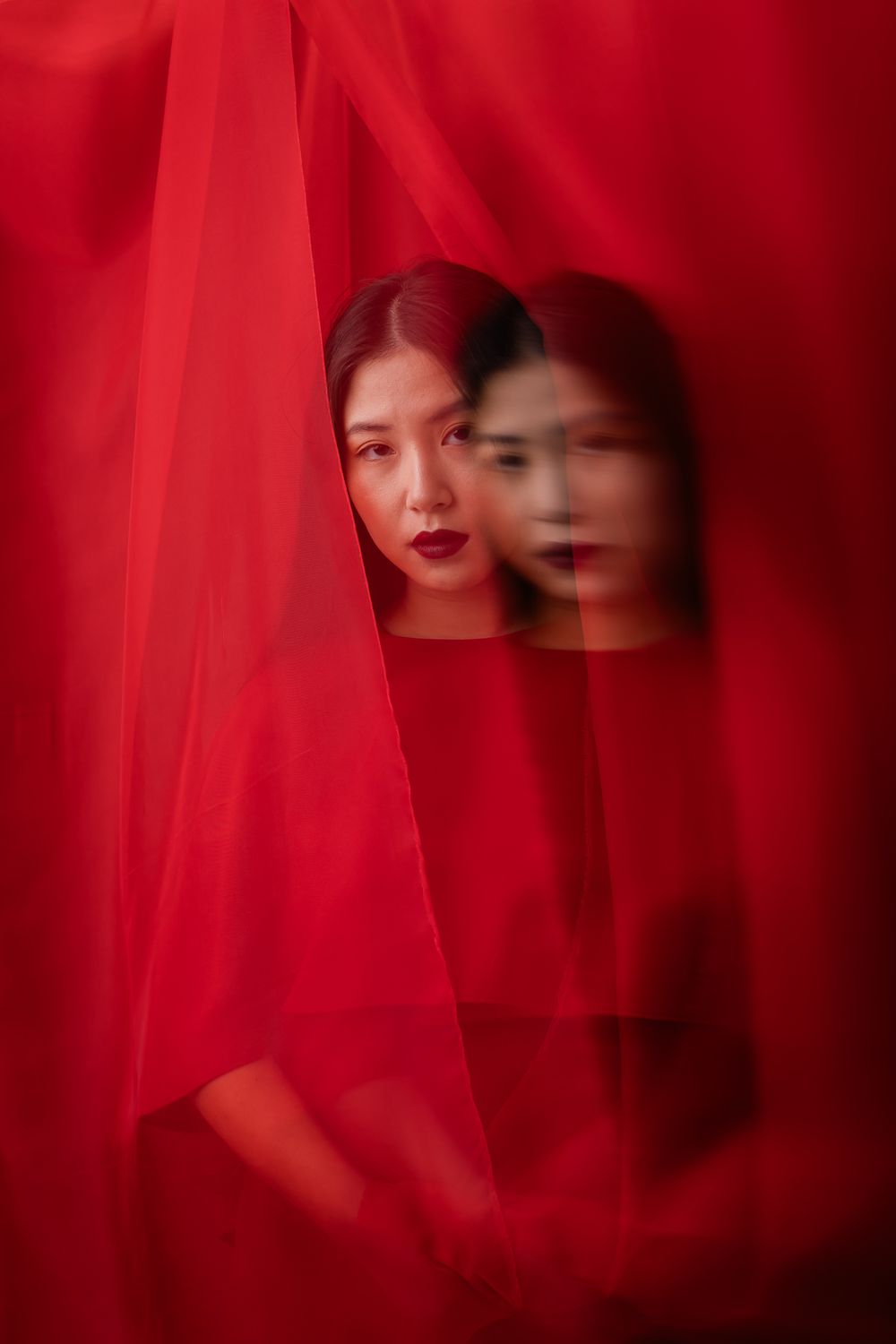 Artistic double exposure portrait behind flowing red fabric creating a ghostly reflection effect.
