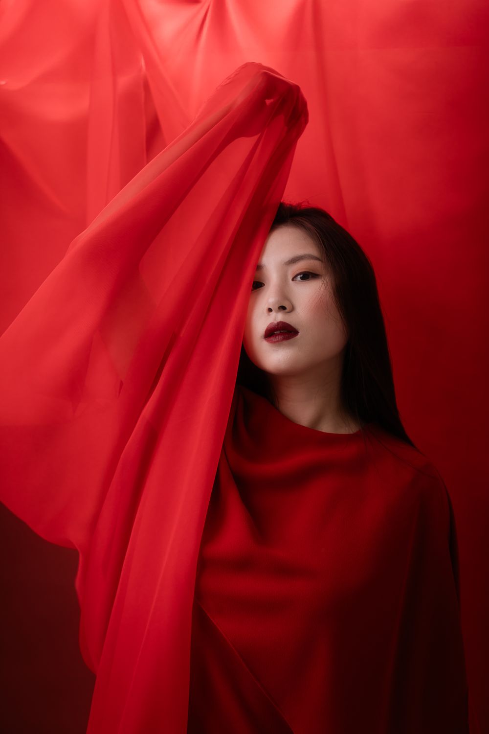 Artistic portrait with red fabric draping and dramatic lighting against deep crimson background.