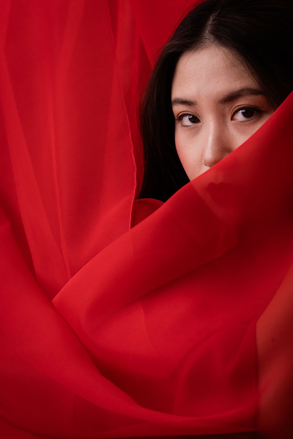 A portrait through flowing red fabric, with dark hair and eyes visible through draped translucent material.
