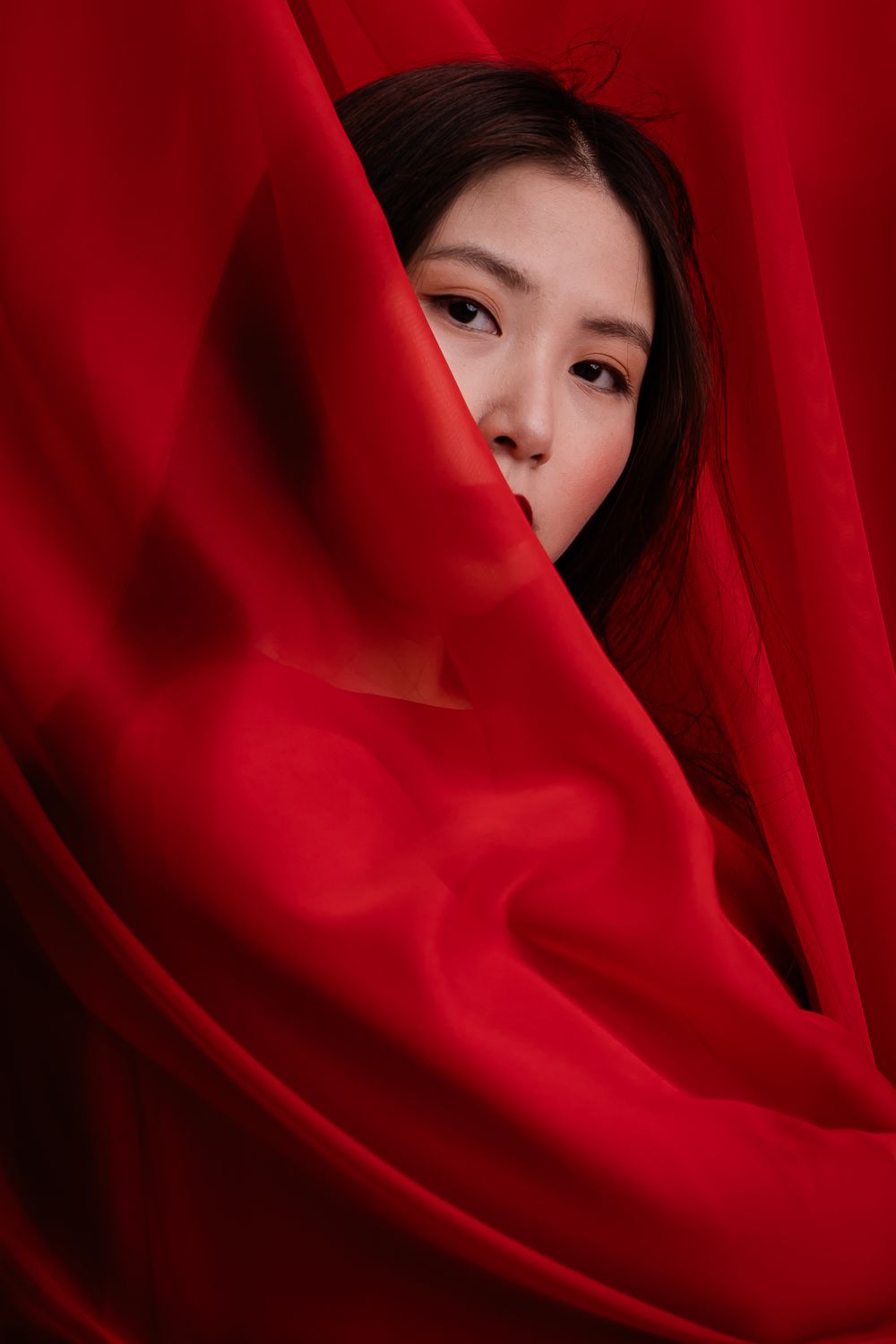 A dramatic portrait through red fabric showing half a face partially hidden in shadow and texture.