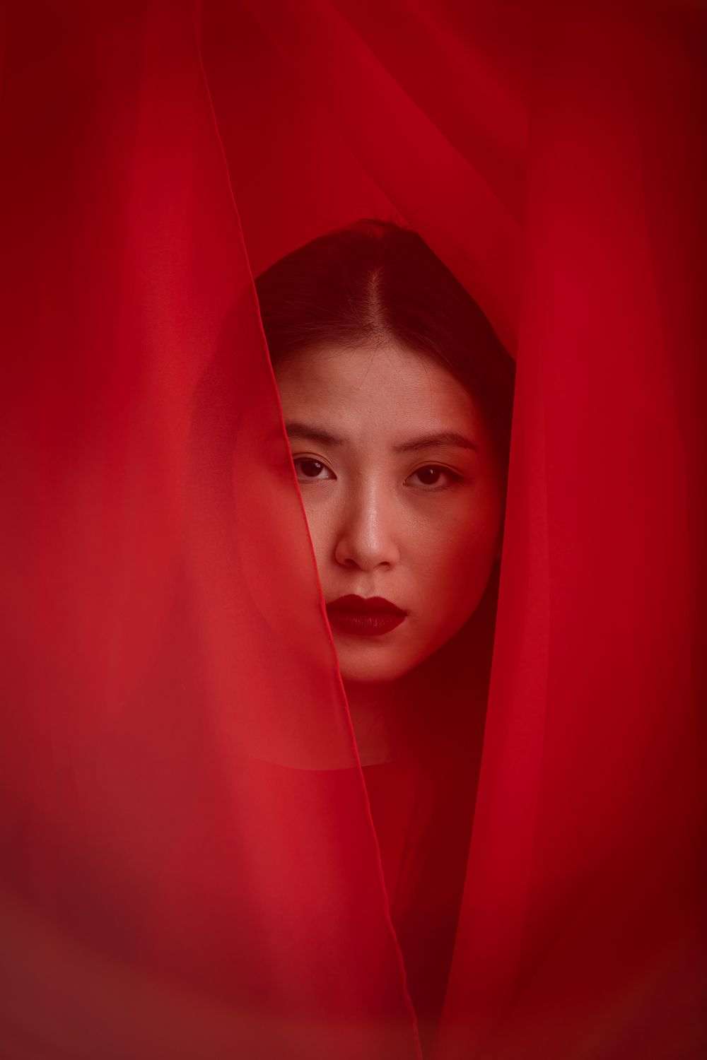 Artistic portrait through red translucent fabric showing dark lipstick and dramatic lighting against crimson background.