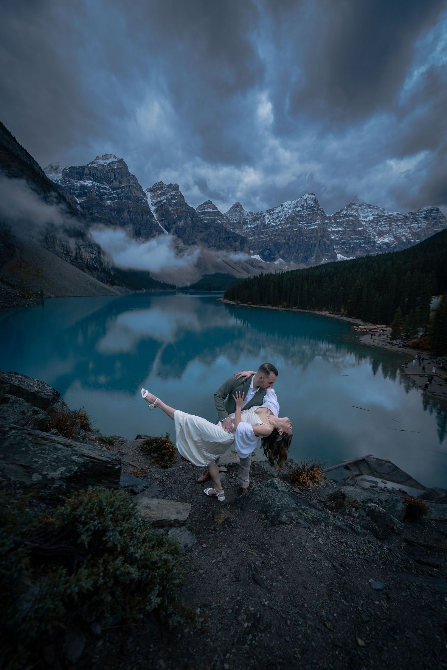 A couple embraces dramatically against the stunning backdrop of Moraine Lake and snow-capped mountains under moody storm clouds.