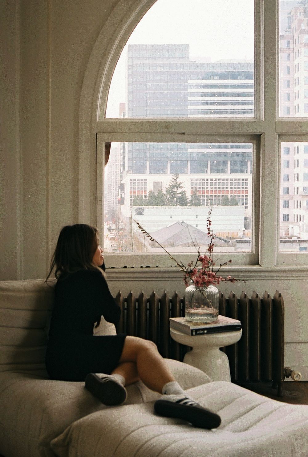 Silhouette by arched window looking out at city skyline with cherry blossoms in vase.