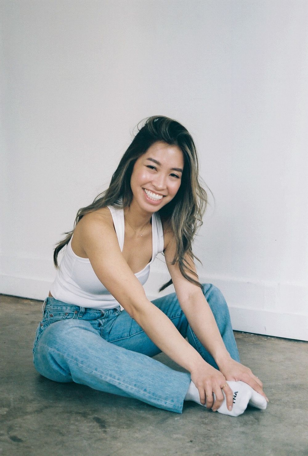 Person sitting casually on floor wearing white tank top and blue jeans in minimalist setting.