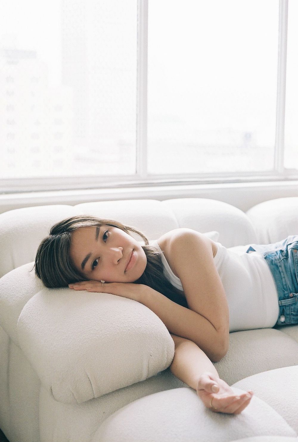 Person in white tank top and jeans lounging on white couch in bright room.
