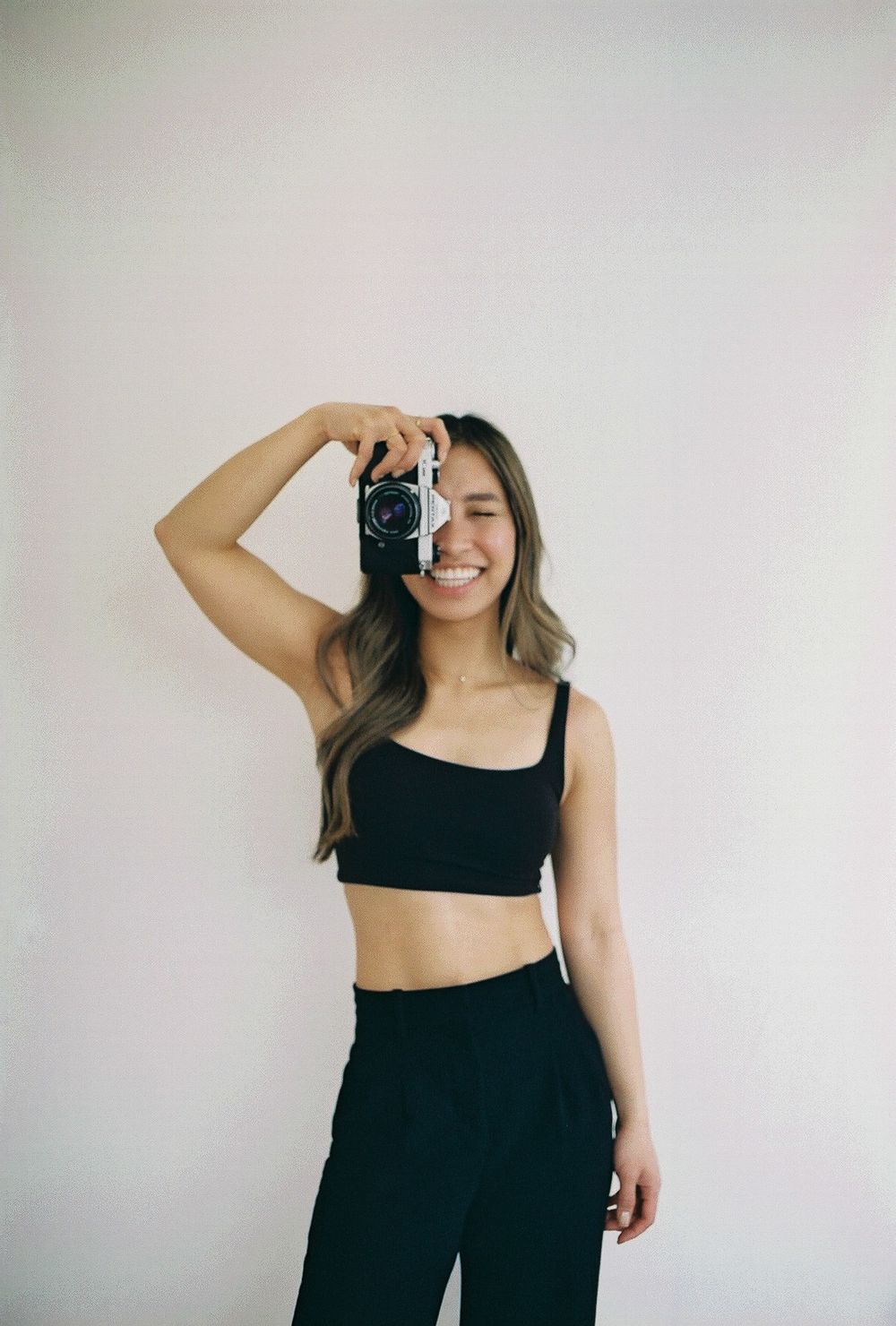 Person taking a photo with a vintage camera while wearing black crop top and pants against white wall.