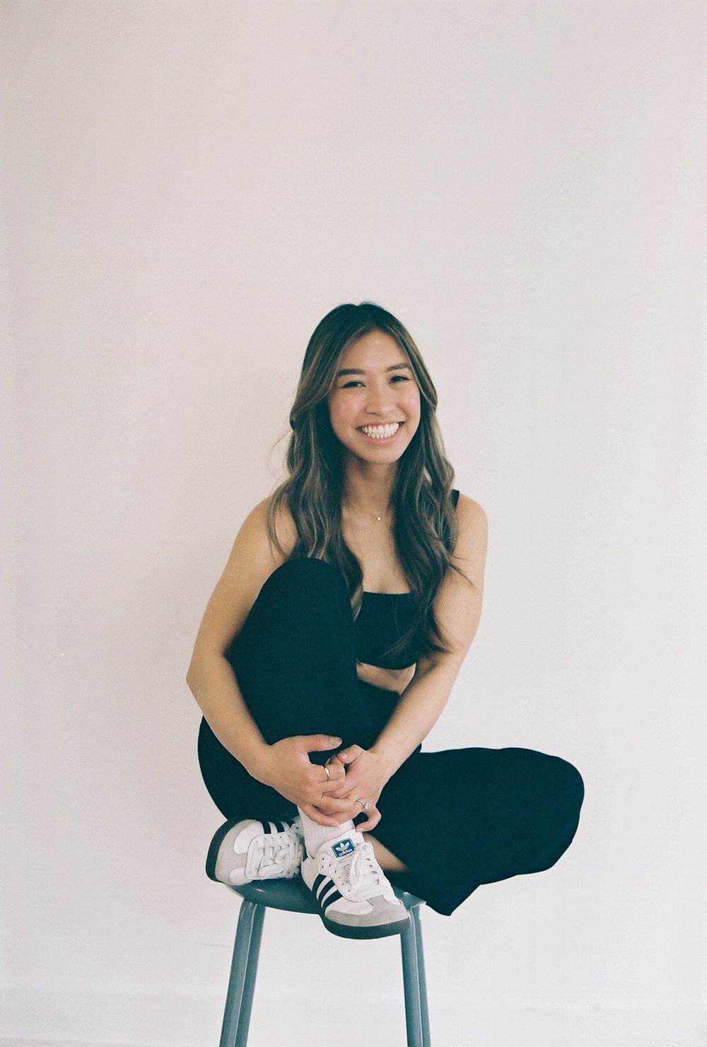 Person in black outfit sitting cross-legged on stool against white background.
