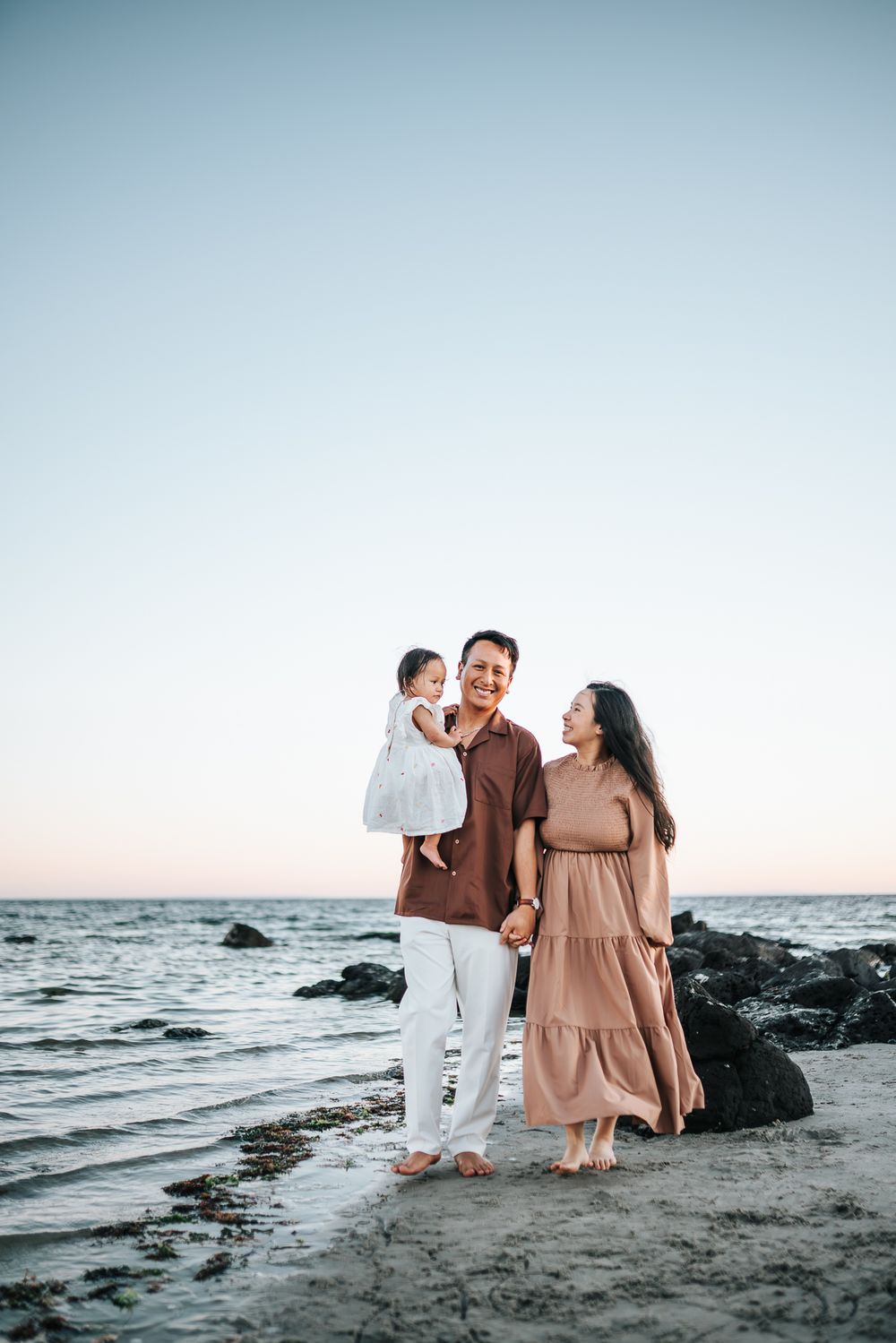 A happy family of three poses on a sandy beach at sunset, with parents holding their baby girl near rocky shores.