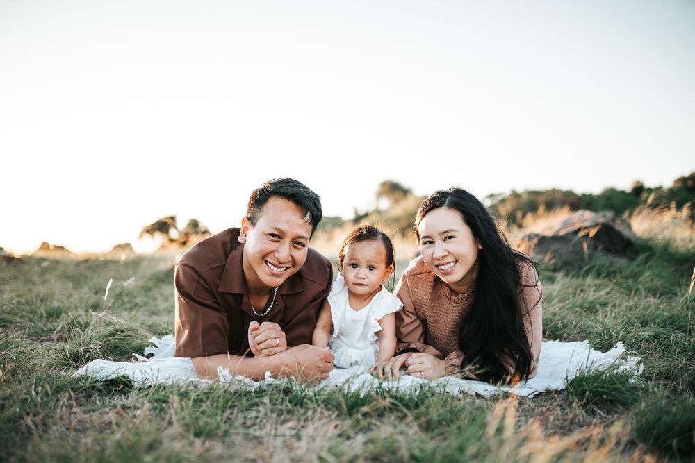 Happy Asian family with baby lying on white blanket in green field during golden hour sunset.