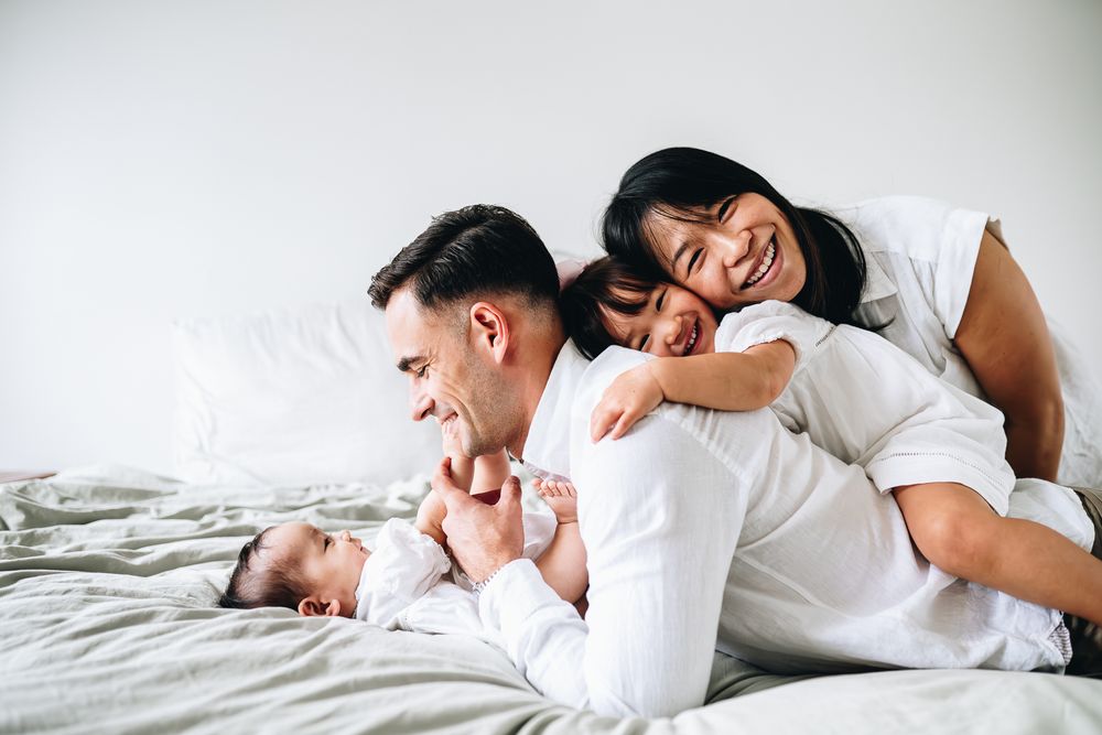 Happy family enjoying playtime together on a white bed wearing matching casual white outfits taken by Melbourne family photographer.