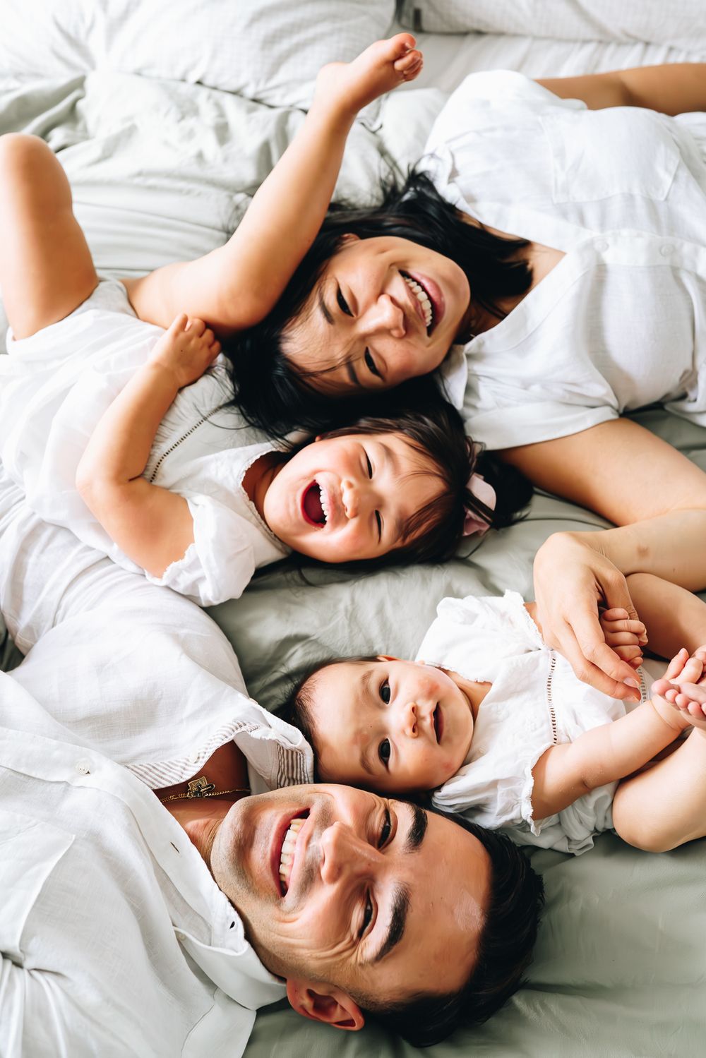 Happy family wearing white clothes lying together on white bedding while laughing and having fun taken by Melbourne family photographer.