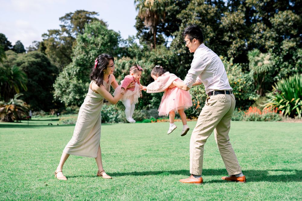 A family plays together on a sunny day, swinging a small child in a pink dress on a green lawn taken by a Melbourne family photographer.