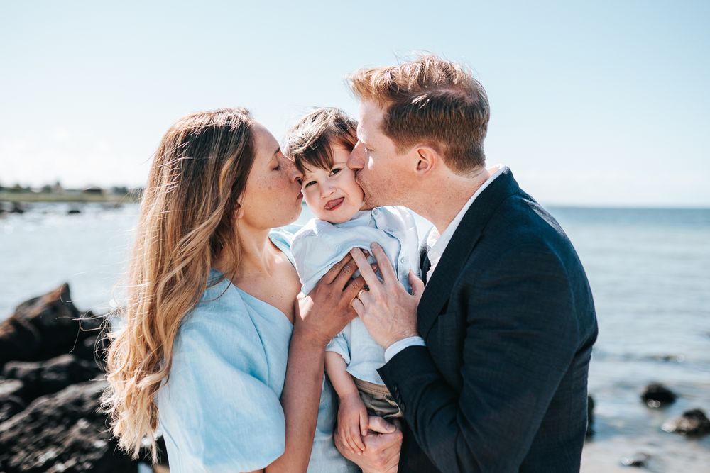 A happy family of three shares kisses on a rocky beach, parents lovingly kissing their toddler son under a clear blue sky.