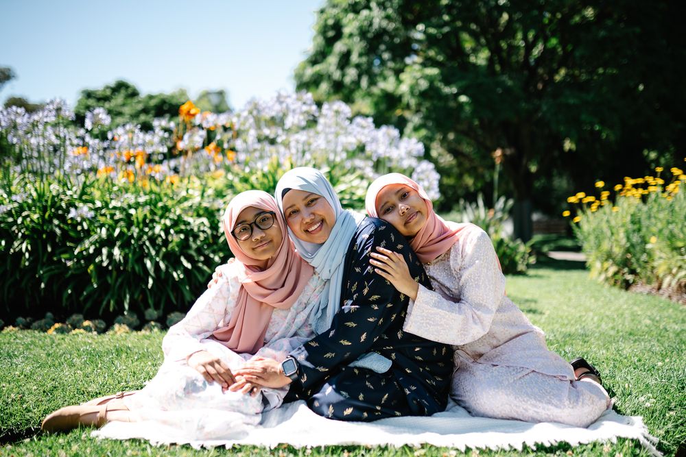Three women wearing hijabs smile and pose together on a blanket in a sunny park with colorful flowers.