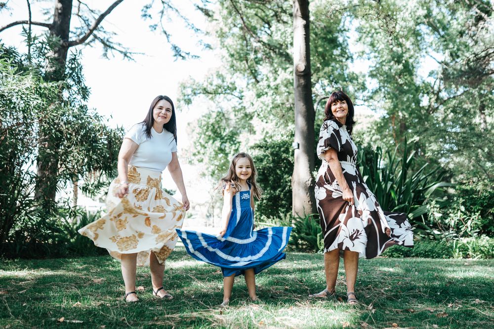 Two women and a girl twirl in floral skirts in a sunny green park, smiling joyfully together.