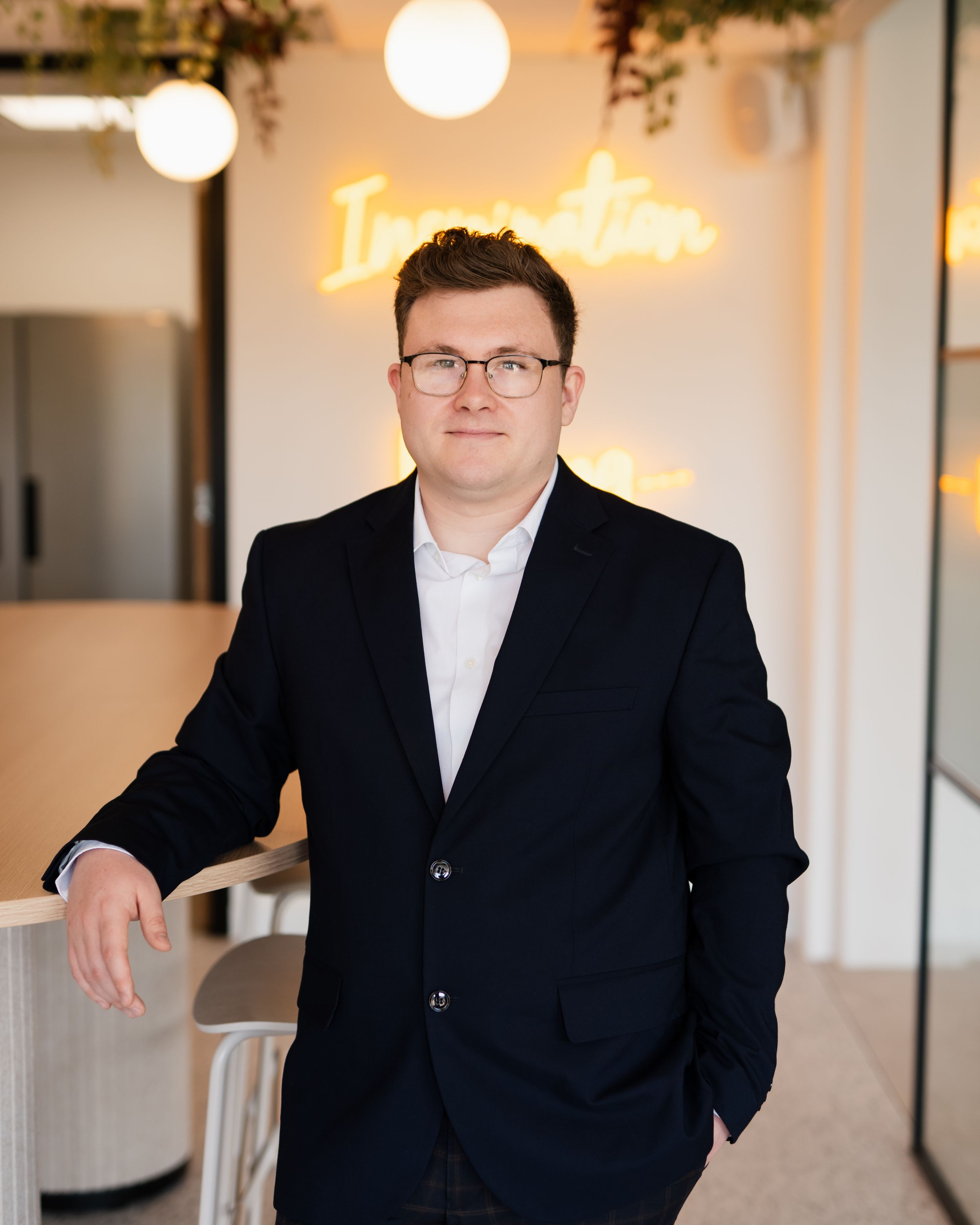 Professional business portrait of person in black suit and white shirt standing in modern office interior with pendant lights.