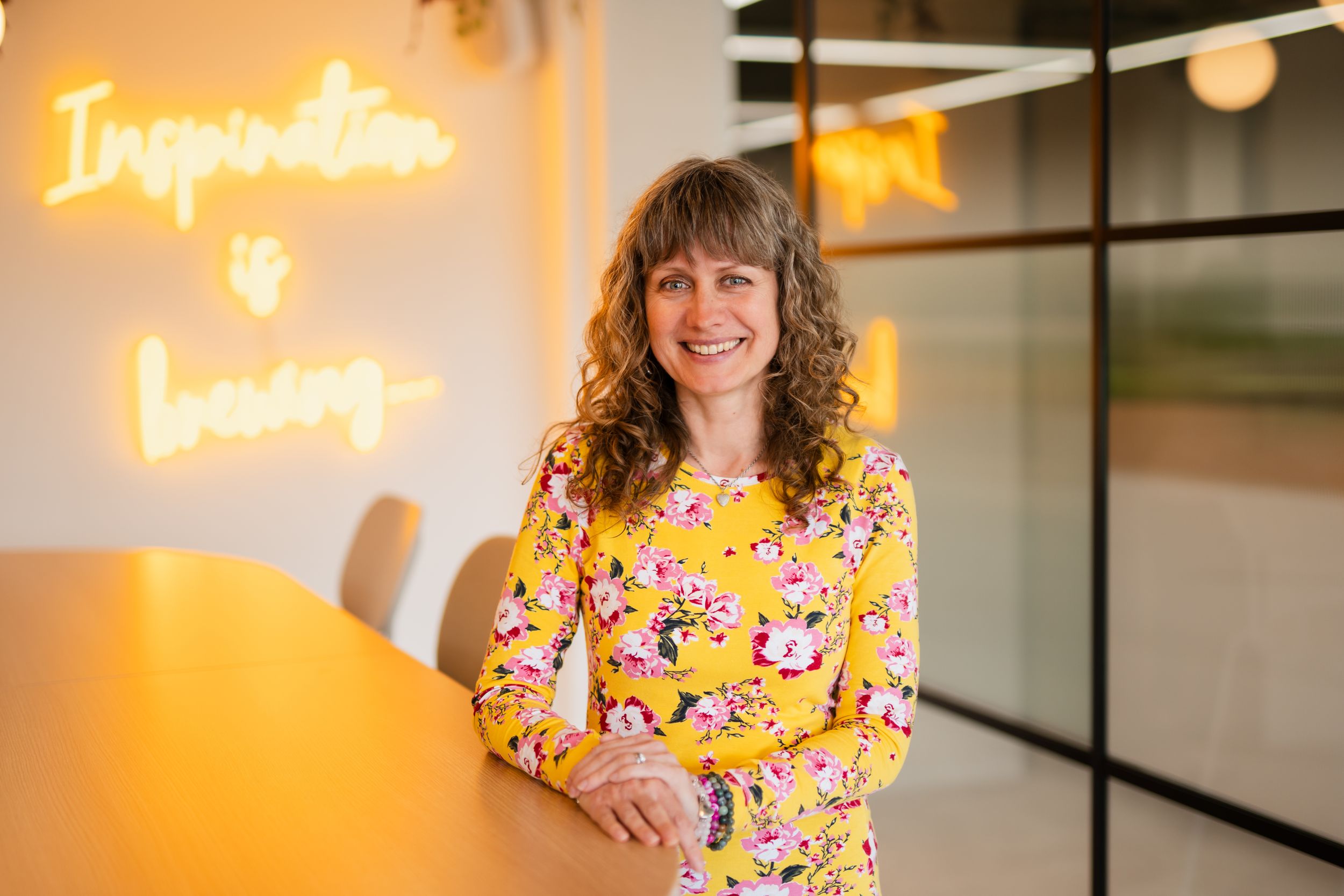 Professional stands in front of illuminated neon sign wearing vibrant yellow floral dress in modern office setting.