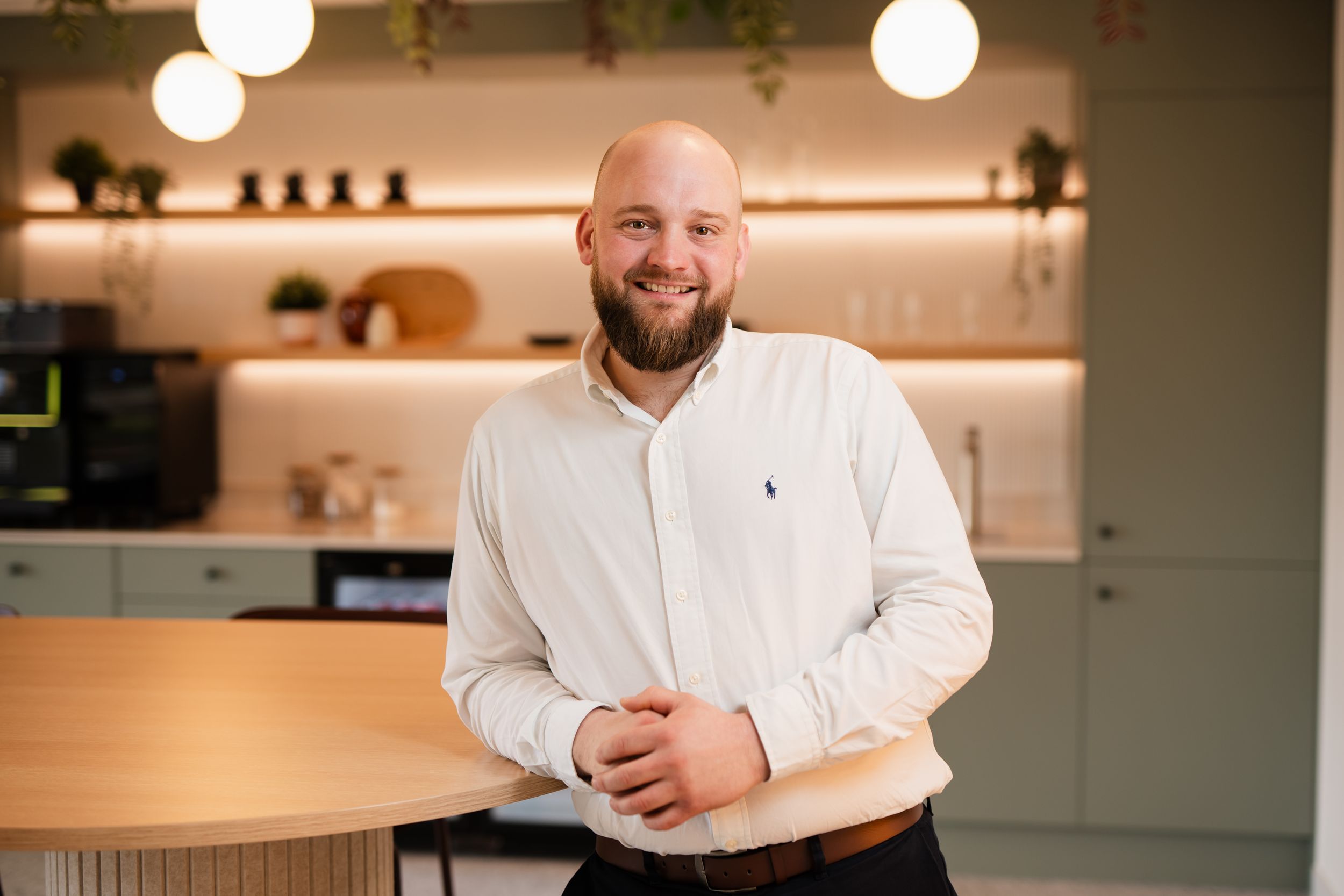 A person wearing a white button-down shirt stands smiling in a modern kitchen with wooden counters and pendant lights.
