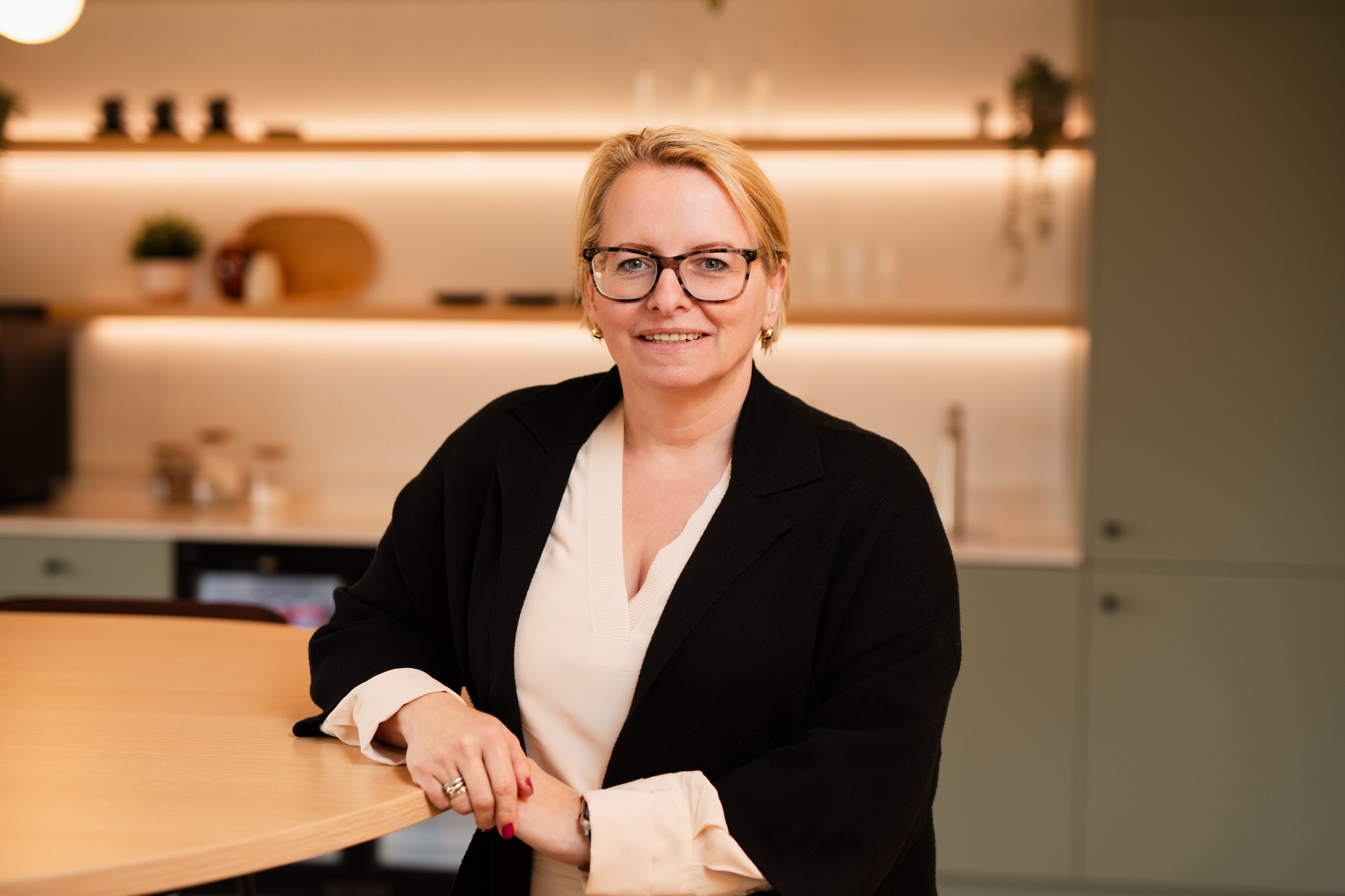 Professional wearing glasses and black cardigan smiles while seated at wooden table in modern minimalist kitchen setting.