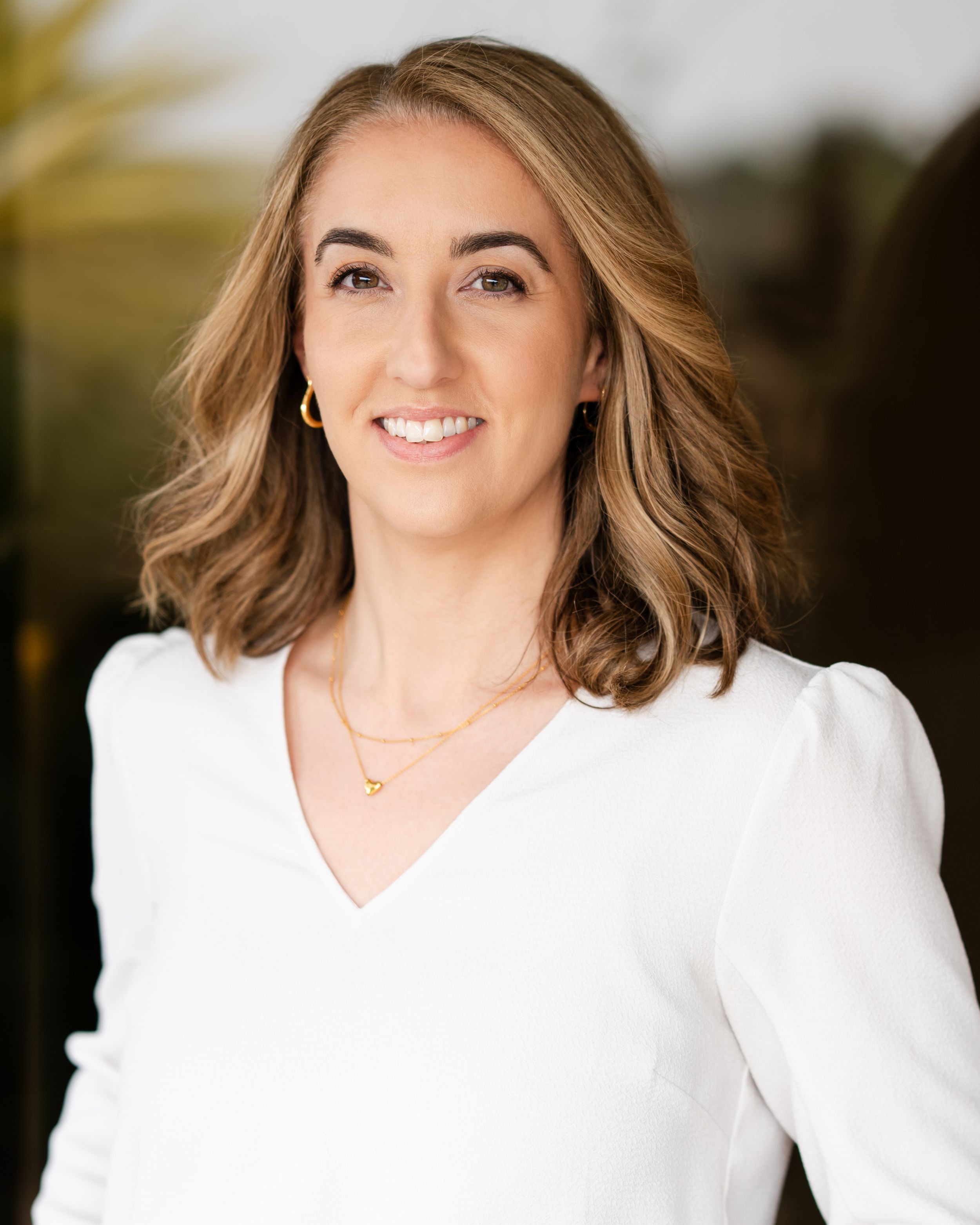 Professional headshot of someone in a white v-neck top with wavy brown hair and delicate necklace against blurred background.