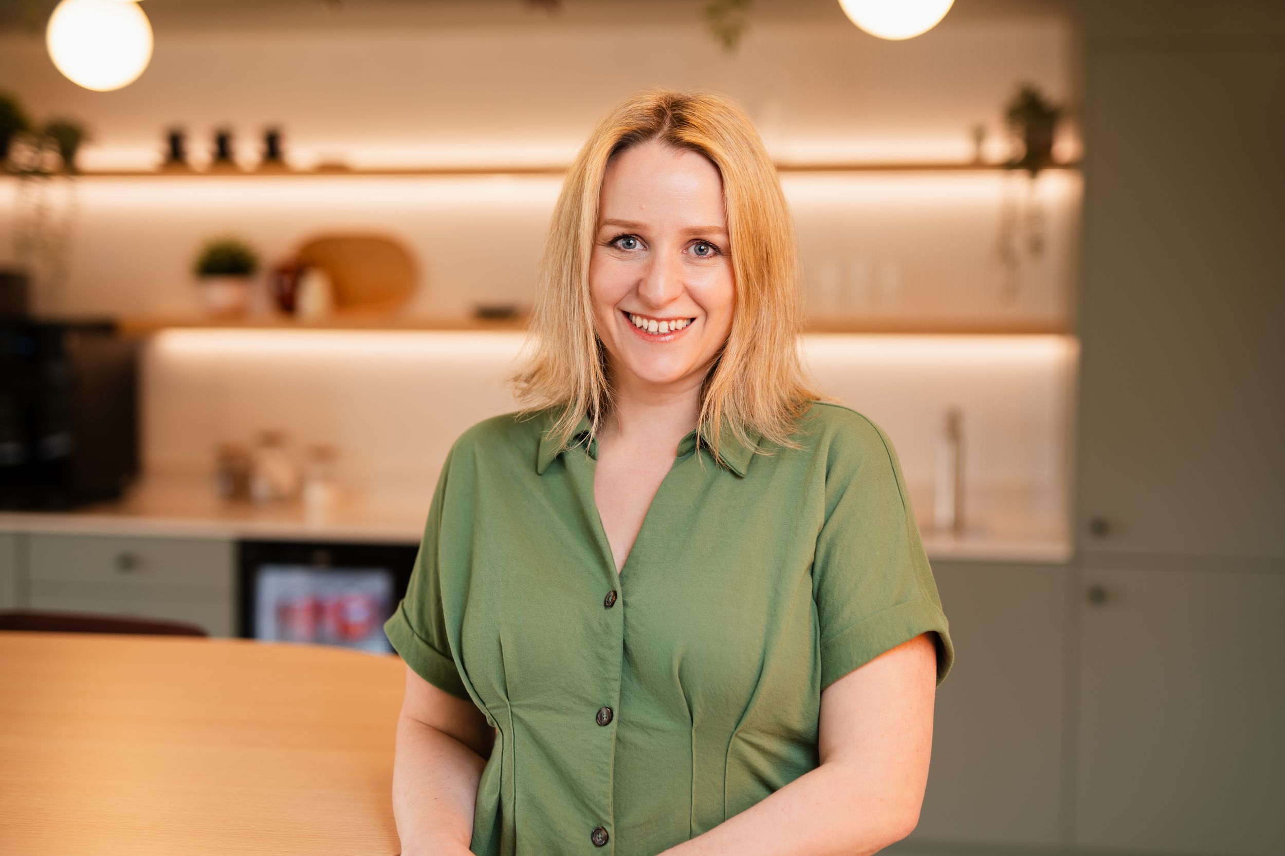 A person wearing a green button-up shirt smiles warmly in a modern kitchen with white shelving and soft lighting.