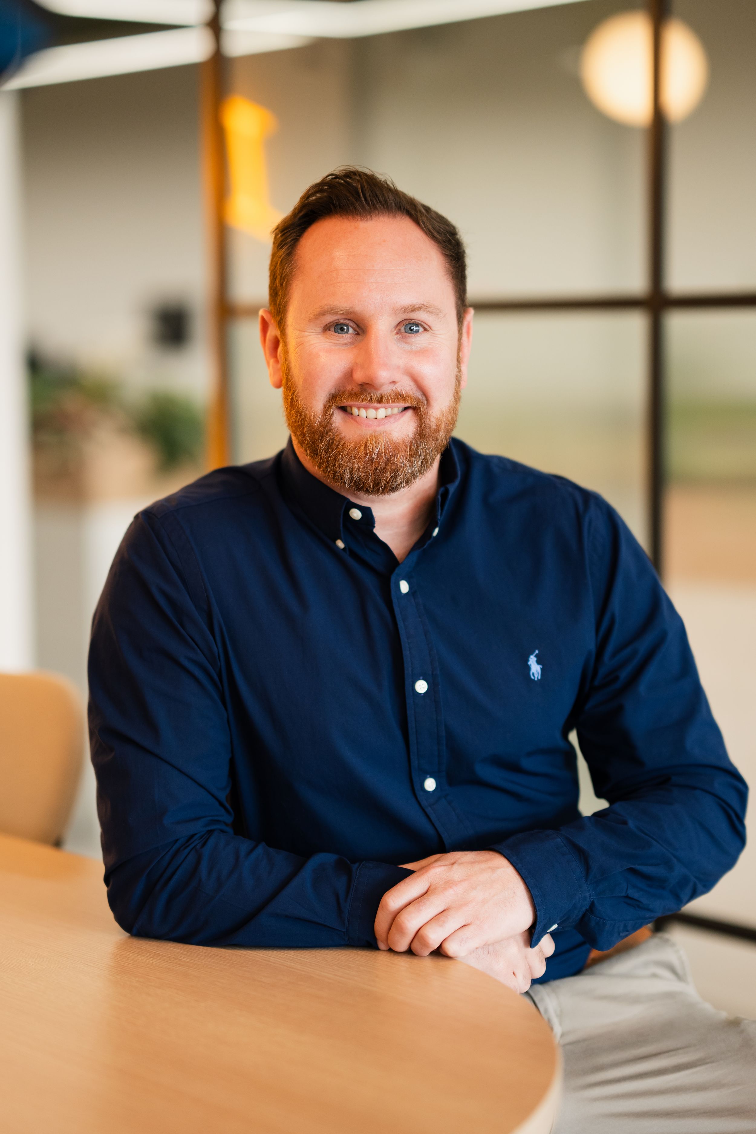 Professional headshot in office setting showing a person in navy button-down shirt seated at wooden table with warm lighting.